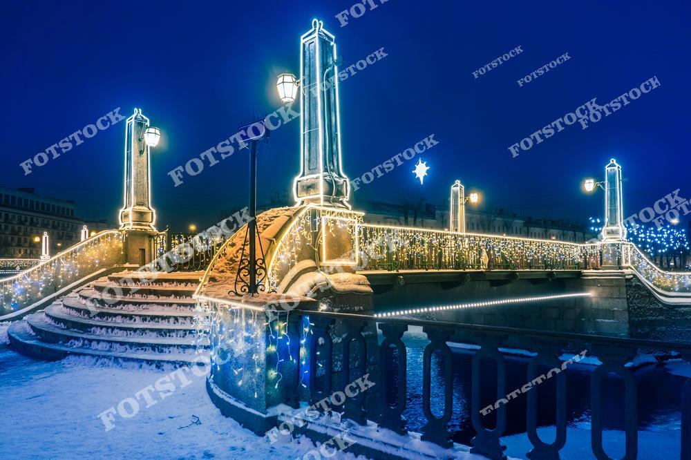 На фотографии изображен Красногвардейский мост, ярко украшенный праздничными гирляндами и световыми конструкциями. Лестницы и перила моста сияют теплым светом, создавая волшебную атмосферу зимнего Санкт-Петербурга. Ночное освещение подчеркивает детали архитектуры моста и отражения огней на заснеженном покрытии. Этот мост является ярким элементом городского ландшафта и привлекает внимание своей элегантностью.